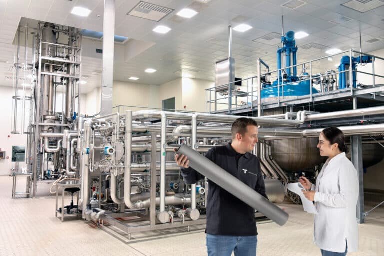 Dos personas en una planta de producción de alimentos revisando el material de aislamiento técnico ThermaSmart Pro para tuberías, rodeadas de equipos de acero inoxidable.